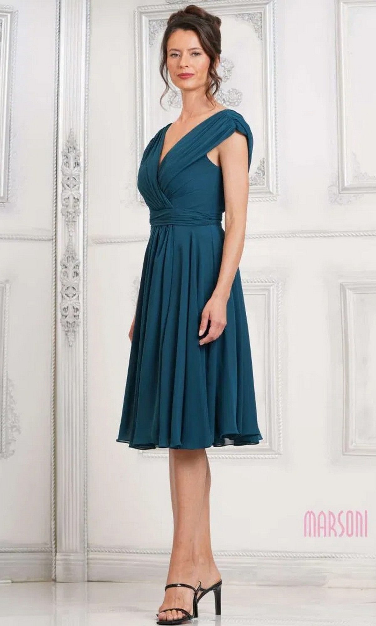 A woman in a knee-length, deep green chiffon Marsoni M251S semi-formal dress with a v-neckline and cap sleeves stands before a white ornate wall, wearing black heeled sandals.